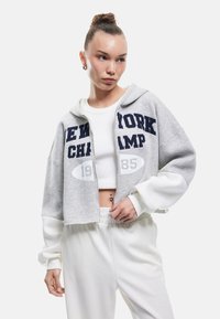 Gekürzter grauer Hoodie mit weißen Ärmeln, der den Text "NEW YORK CHAMP" und "1985" auf der Vorderseite trägt. Kombiniert mit weißen, hochgeschnittenen Hosen.