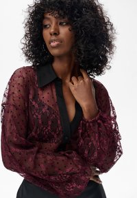Bordowa przezroczysta bluzka z czarnym kołnierzem i zapięciem na guziki, zdobiona kwiatowymi wzorami z koronki oraz falbaniastymi rękawami z kropkowanymi akcentami.