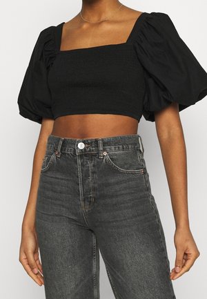Kobieta ubrana w czarny marszczony krótki top z bufkowymi rękawami oraz wysokim stanem, wyblakłe czarne dżinsy, pokazana od szyi do bioder.