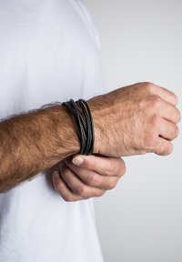 Melnas ādas aproce ar vairākiem pīts pavedieniem, teksturētu virsmu, uz plaukstas ar redzamām roku matiem, uz vienkārša fona.
