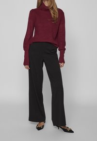 Femme portant un pull en maille bordeaux, un pantalon noir à jambes larges et des escarpins noirs à bout pointu avec un détail chaîne dor ée, sur un fond gris uni.
