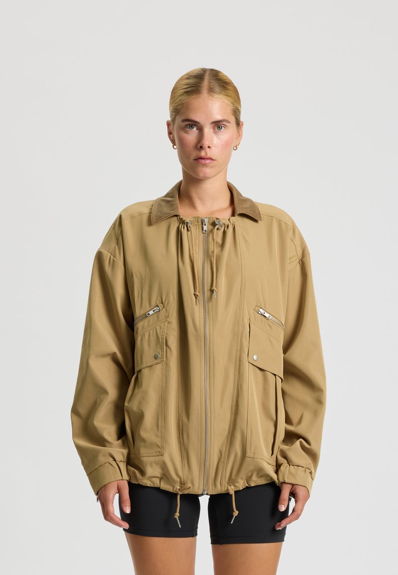 Beige, übergroße Jacke mit einem Kordelkragen, Reißverschluss vorne und zwei großen Seitentaschen. Glattes Material mit einem subtilen Glanz.