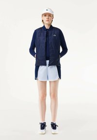 Chaleco acolchado azul marino sobre una camiseta de manga larga azul marino, combinado con pantalones cortos azul claro y zapatillas blancas. Una visera blanca presenta un logotipo verde.