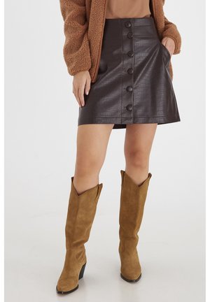 Kvinne iført en brun imitert skinn skjørt med knapper, tan sued støvler med cowboydesign, og en brun teddy-jakke med lommer.