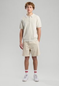 Kerman lyhyet hihat polo-paita vetoketjulla, yhdistetty beigenvärisiin shortseihin. Asua täydentävät valkoiset tennarit ja punaraitaiset sukat.