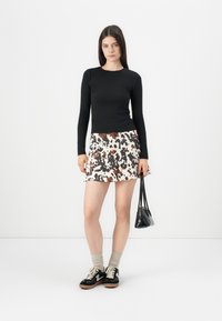 Giovane donna che indossa un top nero a maniche lunghe, una gonna corta con stampa animalier, calze beige e sneakers nere, mentre tiene una piccola borsa nera, in piedi davanti a uno sfondo bianco.