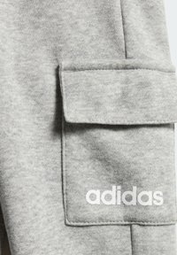 Približni prikaz sive tkanine džepa s preklopom i bijelim logotipom "adidas" otisnutim u donjem desnom kutu.