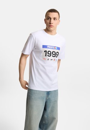 Młody mężczyzna w białej koszulce z nadrukiem "PROSTO" i "1999", w połączeniu z jasnoniebieskimi szerokimi dżinsami, stoi i patrzy w prawo.