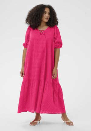 Kobieta z kręconymi włosami w jasnoróżowej, luźnej sukience midi z bufkami na rękawach i niskimi sandałami na obcasie, stojąca na tle jednolitego tła.