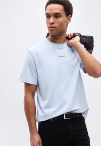 Ανοιχτό μπλε βαμβακερό t-shirt με κοντά μανίκια, με μικρό μαύρο λογότυπο "Calvin Klein" στο στήθος. Φορώντας το με μαύρο παντελόνι και μαύρη τσάντα.