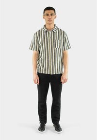 Camicia a maniche corte con righe verticali verdi, beige e blu; colletto a bottone, taschino singolo; abbinata a pantaloni e scarpe nere.