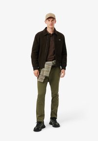 Giacca bomber marrone scuro, polo scura, pantaloni verde oliva, sciarpa grigia a righe, sneakers nere, cappellino beige; outfit casual a strati.