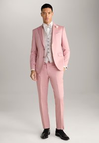 Helles rosa Anzug mit einem taillierten Design, Reverskragen und einer Ein-Knopf-Verschluss; kombiniert mit einer hellgrauen Weste und Fliege. Schwarze Schuhe.