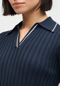 Femme portant un haut en maille côtelée bleu marine avec un col en V fendu comportant des accents de bordure blanche.