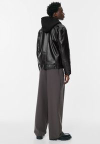 Mladić okrenut bočno nosi crnu kožnu jaknu preko crne dukserice, sive široke hlače i crne cipele.