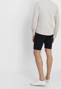 Camisa beige clara de lino con mangas remangadas, combinada con pantalones cortos negros. Modelo con zapatillas blancas, visto desde atrás.