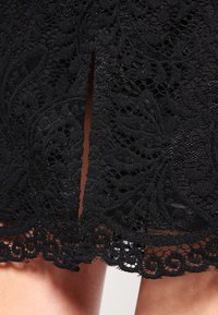 Jupe en dentelle noire avec des motifs floraux délicats, une longueur mi-longue avec un bord festonné et une fente centrale pour une touche supplémentaire.