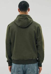 Mand set fra bagenden iført en olivengrøn hættebluse og blå denimjeans foran en ensartet grå baggrund.