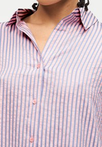 Κοντινό πλάνο ατόμου που φοράει πουκάμισο με κάθετες ρίγες σε ροζ και μπλε χρώμα, με γιακά και λαιμόκοψη σε σχήμα V.