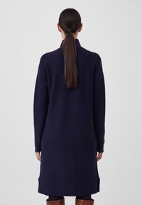 Long pull en maille bleu marine avec un col montant et des épaules tombantes, présentant une coupe droite, des fentes latérales et une texture douce.