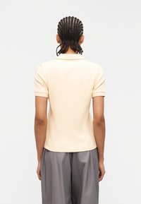 Άτομο με πλεγμένα μαλλιά φοράει κοντομάνικο μπεζ πόλο πουκάμισο και φαρδιά γκρι παντελόνια, στέκεται με την πλάτη προς τα εμάς μπροστά σε απλό φόντο.