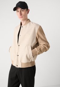 Bežna pletena bomber jakna s tan rukavima, rebrastim manžetama in srebrnimi gumbi. Usklajena z črno majico in črnimi hlačami. Enostavna ozadja.
