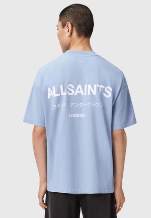Persoon met kort haar die een lichtblauw T-shirt draagt met "ALLSAINTS" en Japanse tekst boven "LONDON" gedrukt op de achterkant.