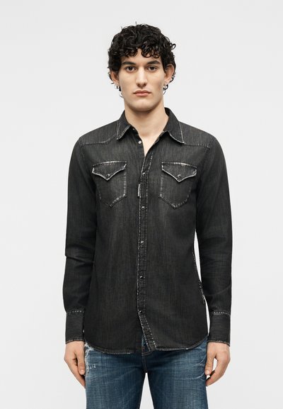 Νέος άνδρας με σγουρά σκούρα μαλλιά, φοράει μαύρο πουκάμισο τζιν με κουμπιά και δύο τσέπες στο στήθος, καθώς και μπλε τζιν, στέκεται μπροστά σε απλό φόντο.