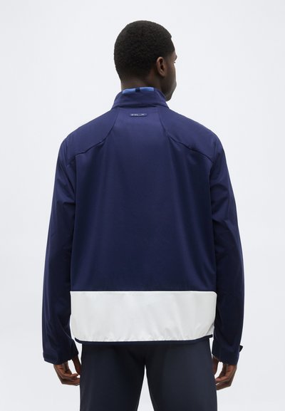 Hombre visto desde atrás, vistiendo una chaqueta azul marino con un panel horizontal blanco en la parte inferior, de pie frente a un fondo liso.