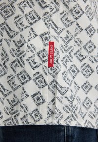 Witte stof met een blauw geometrisch patroon, met een rood label met de tekst "JOOP! JEANS" nabij een naad. Het ontwerp is ingewikkeld met een texturaal uiterlijk.