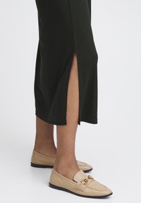 Mörkgrön knälång klänning med sidoklipp, tillsammans med beige loafers med guldtonad detalj och en slät lädertextur.