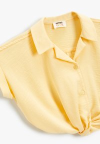 Camisa amarilla de cuello con mangas cortas, con tapeta de botones en la parte frontal, detalle de dobladillo anudado, tela de textura suave. Etiqueta de la marca visible en el cuello.