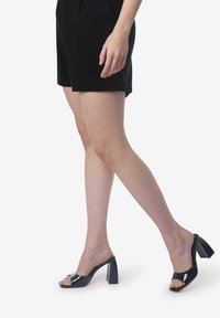 Donna che indossa mule aperti con tacco alto neri e pantaloni shorts neri, camminando con una gamba estesa in avanti.