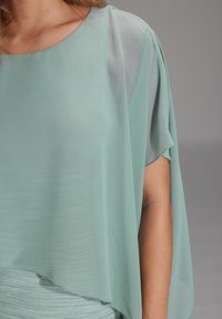 Haut léger en teal, ample, avec des manches transparentes et un col rond. Texture douce et lisse avec des rayures irisées horizontales à l'ourlet.