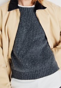 Beżowa kurtka zapinana na zamek z kołnierzem z sztruksu, noszona na ciemnoszarym prążkowanym swetrze. Miękkie tekstury i dopasowany krój podkreślają warstwowanie.