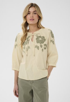 Femme portant un chemisier crème avec broderie florale verte aux épaules et aux manches, associée à un pantalon vert olive, debout devant un fond gris.