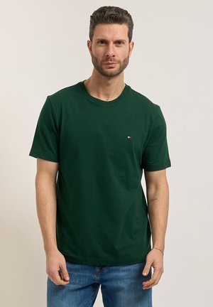 Uomo con capelli corti e ricci e barba, che indossa una t-shirt a maniche corte verde scuro e jeans blu, in piedi contro uno sfondo chiaro e uniforme.