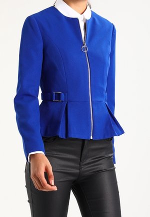 Kobieta ubrana w dopasowaną królewską niebieską kurtkę na zamek z bocznymi klamrami i plisowanym dołem, w połączeniu z czarnymi skórzanymi spodniami.