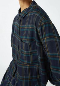Camicia a quadri nei colori blu navy, verde e giallo. Presenta una chiusura con bottoni sul davanti, colletto e tasca laterale. Realizzata in tessuto testurizzato.