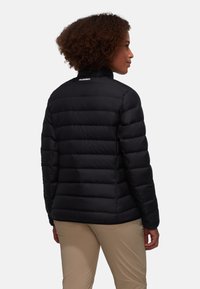 Zwarte pufferjack met horizontaal gestikt patroon, hoge kraag en logo op de achterkant; gecombineerd met lichtbeige broeken.