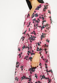 Kvinna i en långärmad rosa blommig klänning med V-ringning och knuten midja, som visar mellansektion och arm mot en enkel ljus bakgrund.