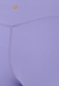 Nahaufnahme eines lavendelfarbenen Stoffs mit detaillierter Naht und einem kleinen goldenen runden Logo nahe dem oberen Rand.