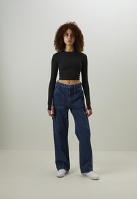 Top crop preto de mangas longas combinado com calças de ganga azuis de perna larga, com grandes bolsos frontais e cintura alta. Ténis brancos completam o visual.