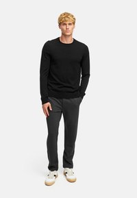 Pull en maille noir à manches longues, associé à un jogging gris foncé et des baskets blanches, avec des accents noirs et beiges.