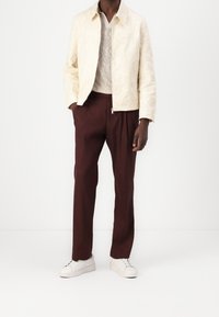 Kremfärgad, broderad bomberjacka lager på lager över en texturerad tröja, kombinerad med mörk vinröda byxor och vita sneakers.