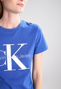 Kvinna iklädd en blå Calvin Klein Jeans T-shirt med stora vita "CK" bokstäver, beskuren från näsan till mitten av överkroppen mot en enfärgad bakgrund.