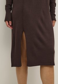 Robe en tricot brun avec une fente sur le côté, des manches longues, des poignets côtelés et une texture lisse. Sa longueur arrive mi-mollet, offrant une coupe décontractée.
