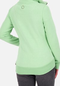 Jersey pulóver verde menta con un alto cuello, puños y dobladillo acanalados, y un detalle de logo circular en la parte posterior del cuello. Textura de tela suave.