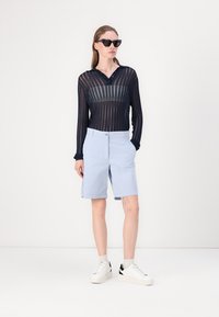 Shorts de culoare albastru deschis cu închidere cu nasture, asortați cu un top lung cu mâneci lungi, transparent, de culoare închisă, cu dungi orizontale, purtându-se cu adidași albi.