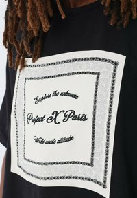 Camiseta de algodón negra con un gran gráfico de paisley en blanco, borde en negro y gris, y el texto "Project X Paris" y "Mundo actitud".